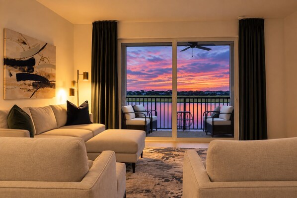 A living room with a sofa, armchairs, and a painting on the wall. There is a balcony with a view of a sunset over a body of water.