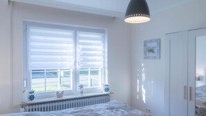 Une chambre à coucher avec un lit, une fenêtre avec des stores, un radiateur, un luminaire au plafond et une armoire.