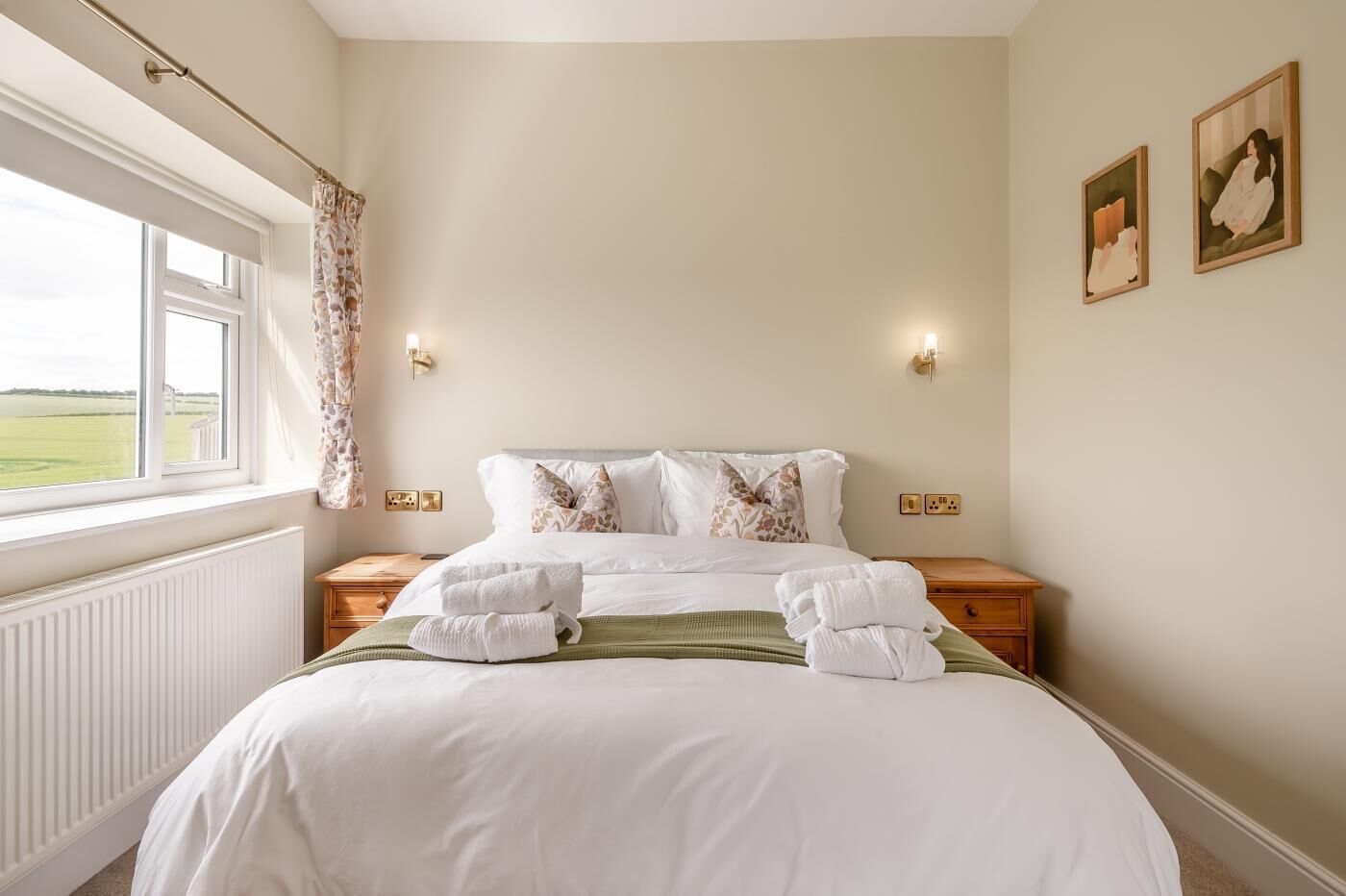 Una cama bien hecha con ropa de cama y almohadas blancas, una ventana con vistas al exterior y dos lámparas fijadas a la pared.