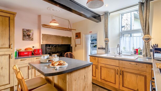 A kitchen with a central island, wooden cabinets, a built-in oven, and a window with curtains. - 3 Bed in Reeth (oc-b42655) (Reeth)