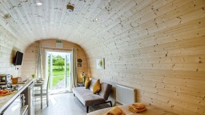 A cozy room with a wooden ceiling, a sofa with cushions, a table with a kettle, and a view of the outdoors.