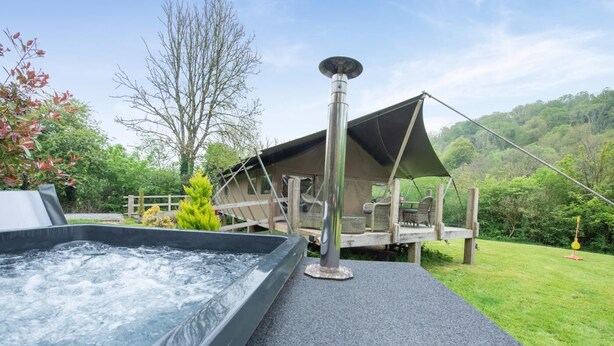 A hot tub adjacent to a wooden deck with a canopy, surrounded by a lush green landscape.