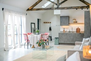 Ein modernes Wohnzimmer mit Glastisch, ein Essbereich mit floralen Stühlen, eine Küche mit grauem Schrank und eine Loft-Decke mit freiliegenden Balken.
