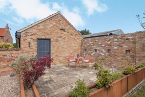 A brick building with a blue door, a small patio with a table and chairs, and a garden area with plants. - 1 Bed in Gembling (oc-k42457) (Gembling)