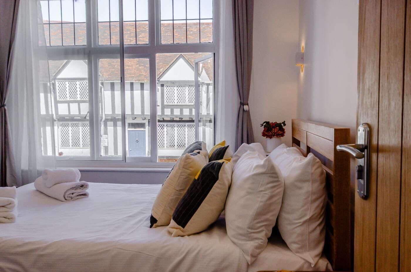 A hotel room with a bed, a window with curtains, a wooden door, and a small vase with flowers.