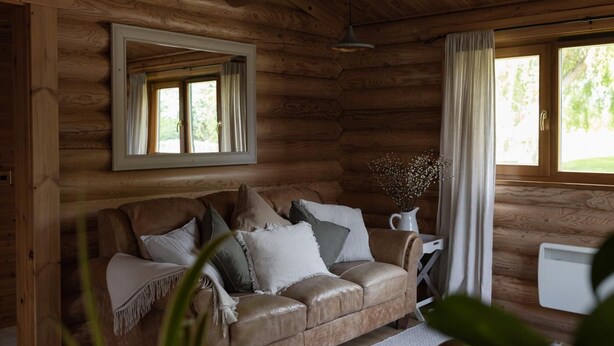 A wooden cabin interior with a leather sofa, a mirror, a window with curtains, and a radiator. - 2 Bed in Cretingham (oc-m42623) (Cretingham)