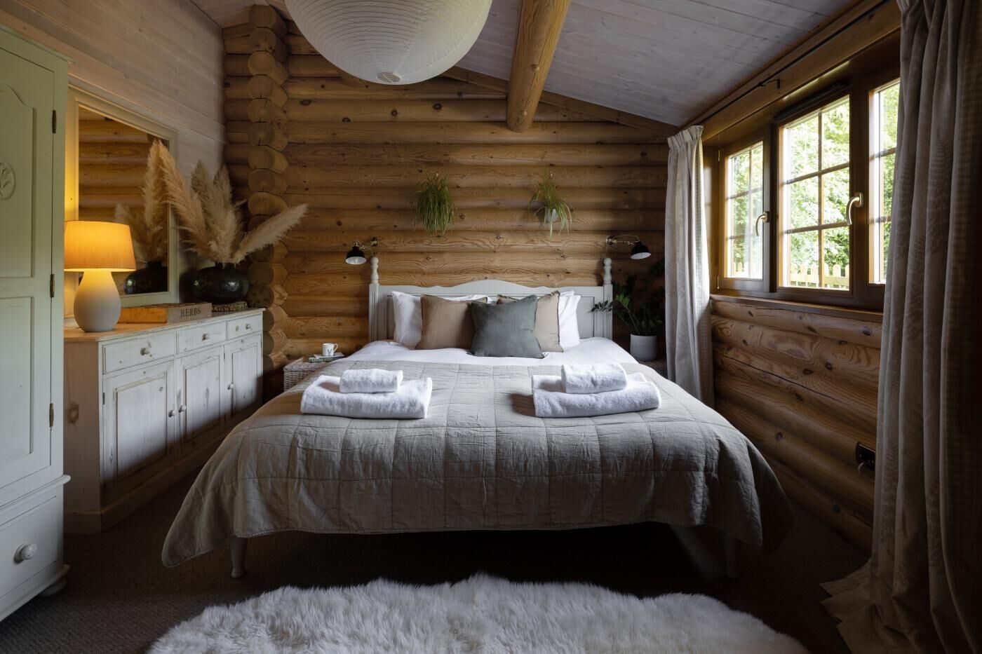 Une chambre à coucher située dans un chalet en rondins, avec un lit, des lampes de chevet, une commode et une fenêtre avec des rideaux.