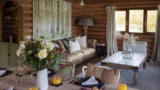 A cozy log cabin interior with a dining area, a sofa, a painting, and a table set for breakfast.