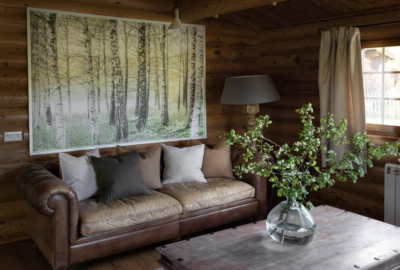 L’intérieur d’un chalet en bois, avec un canapé en cuir, un grand tableau représentant une forêt, une lampe et un vase décoré de plantes vertes.