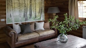 A wooden cabin interior with a leather sofa, a large forest painting, a lamp, and a vase with greenery.