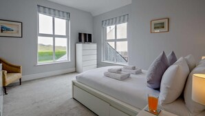 Une chambre à coucher avec un lit, un téléviseur posé sur une commode, une chaise et deux fenêtres avec des rideaux.