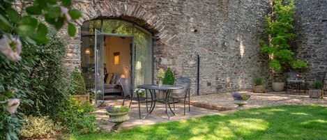 A stone building with a glass door entrance, a patio with chairs, and a grassy garden area.