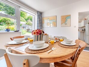 A dining area with a round wooden table, white chairs, and a view of the garden through large windows. - 3 Bed in Tenby (oc-fb189) (Tenby)