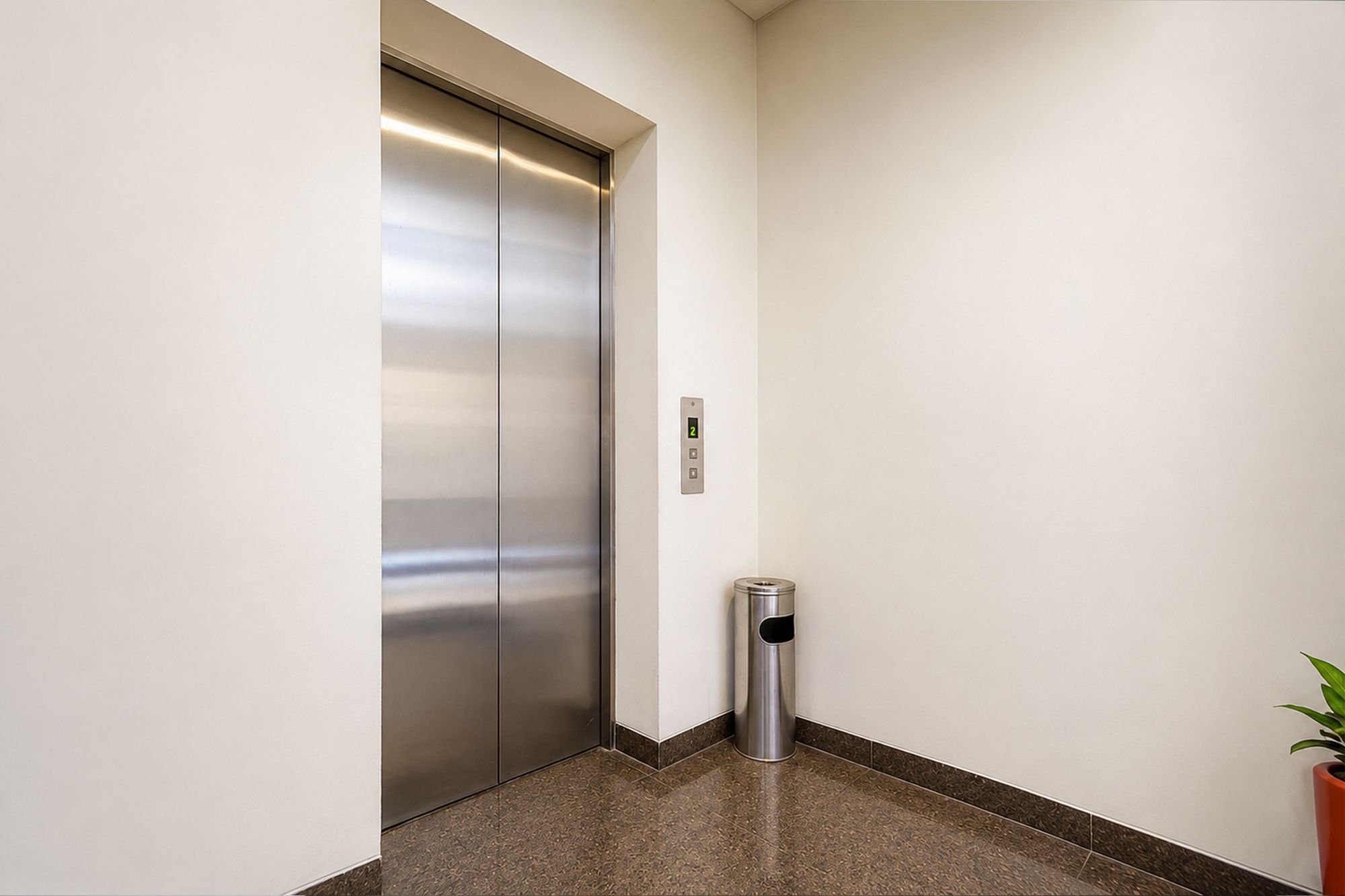 A modern elevator with a digital display, a trash bin, and a potted plant in a hallway.