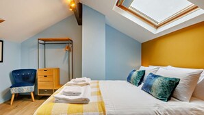 Ein Schlafzimmer mit Dachfenster, einem Bett mit weißer Bettwäsche, einem blauen Sessel, einem Nachttisch aus Holz und einem Kleiderschrank.