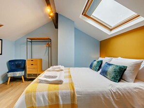 Ein Schlafzimmer mit Dachfenster, einem Bett mit weißer Bettwäsche, einem blauen Sessel, einem Nachttisch aus Holz und einem Kleiderschrank.
