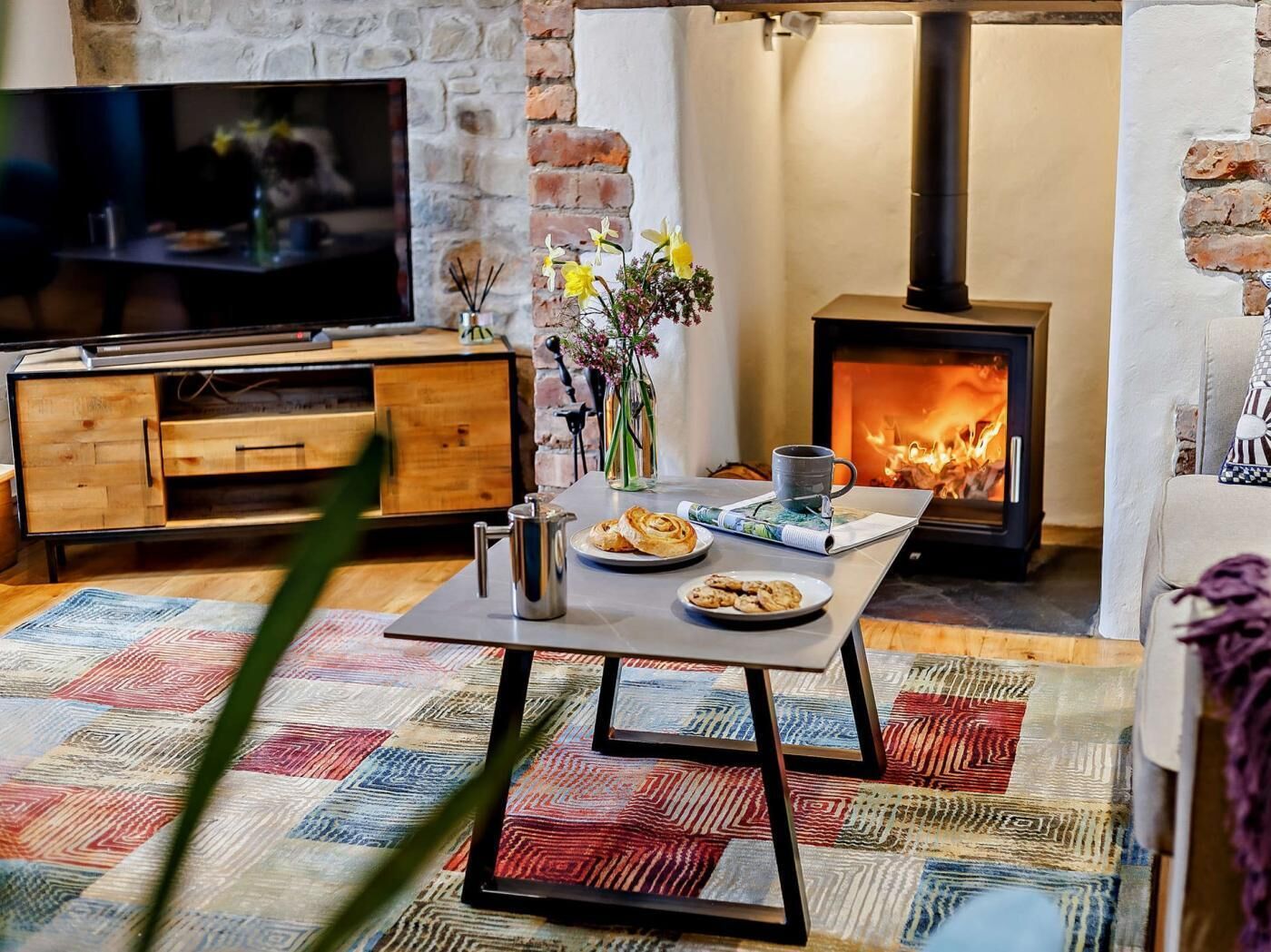 A cozy living room with a television, a table with a mug, cookies, and a vase of flowers, a fireplace, and a stone wall.
