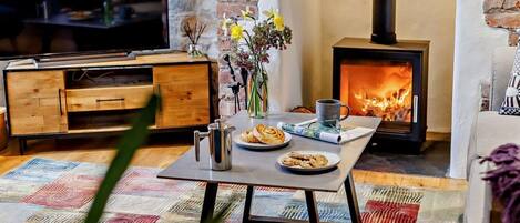 A cozy living room with a television, a table with a mug, cookies, and a vase of flowers, a fireplace, and a stone wall.