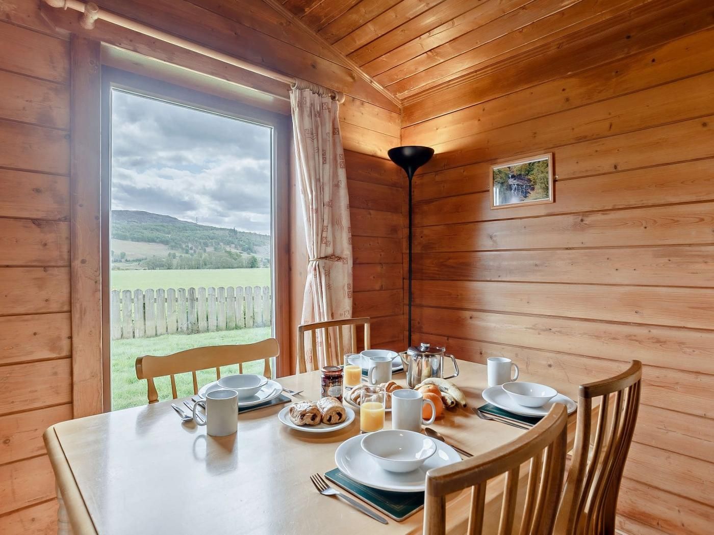 Una baita di legno con un tavolo apparecchiato per la colazione, vista su un paesaggio verde e un quadro appeso al muro.