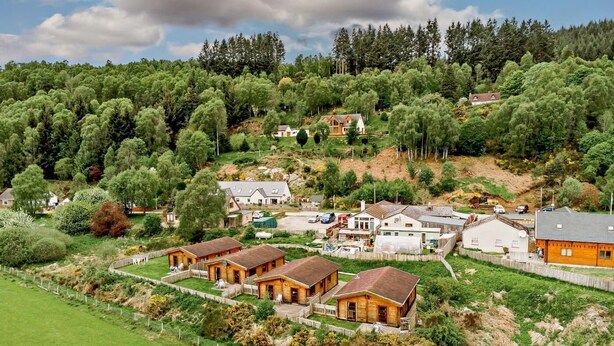 A rural village with several wooden houses, a small road, and a hill with dense trees in the background.