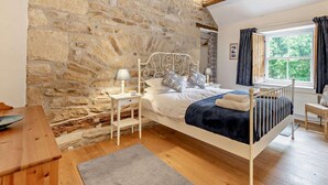 Une chambre avec un mur en pierre, un lit avec un cadre en métal blanc, une table de chevet avec une lampe et une fenêtre avec des rideaux.