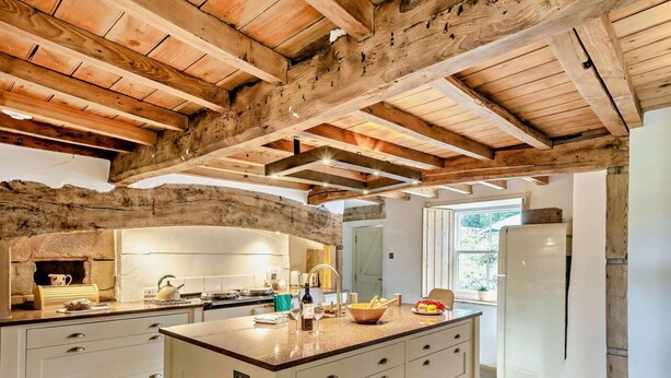 Une cuisine dotée d’un grand îlot central, de poutres en bois, d’un mur en pierre et offrant une vue sur l’extérieur.