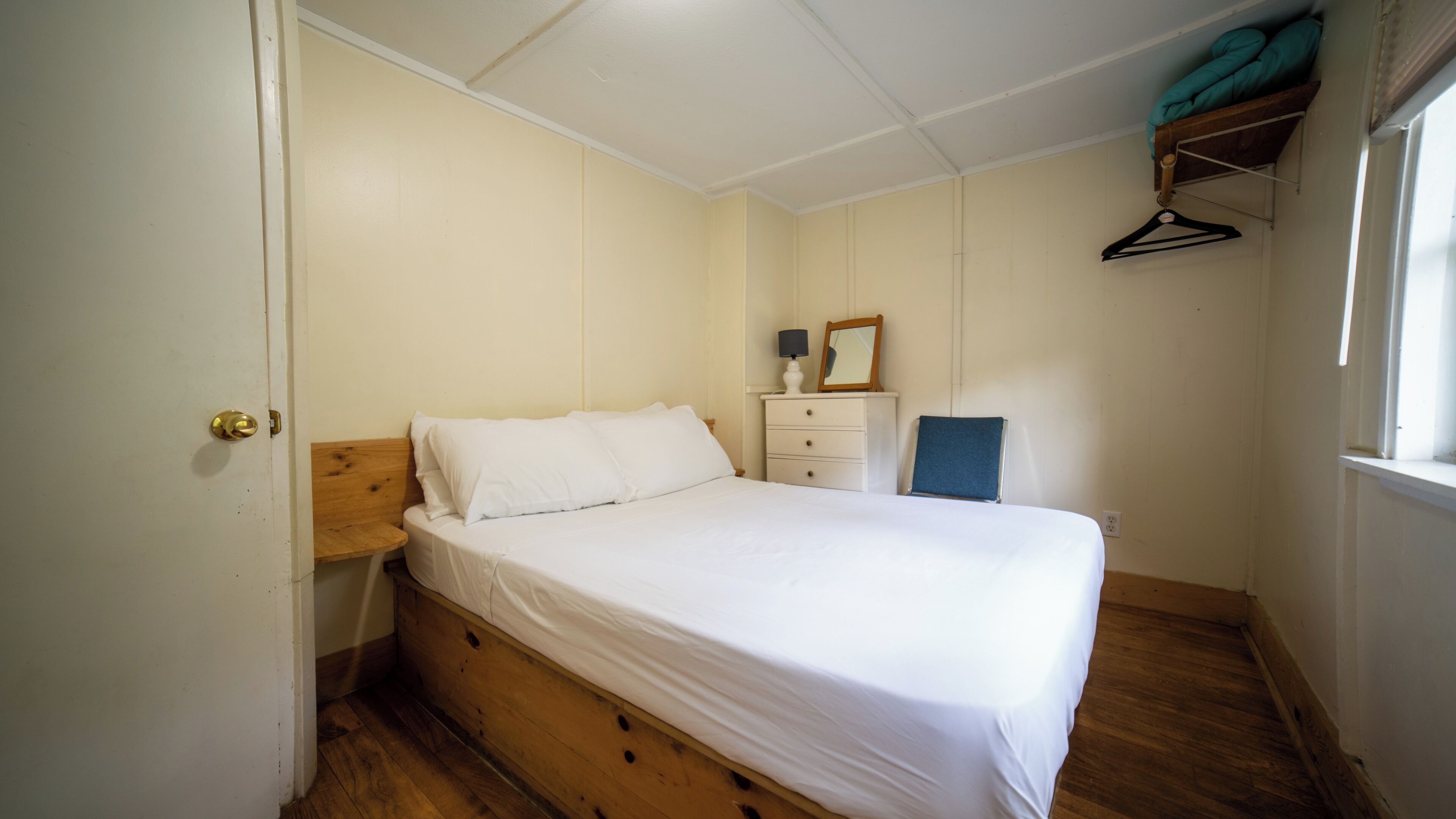 Une chambre à coucher avec un lit en bois, une table de chevet, une commode, une chaise et une fenêtre donnant sur l’extérieur.