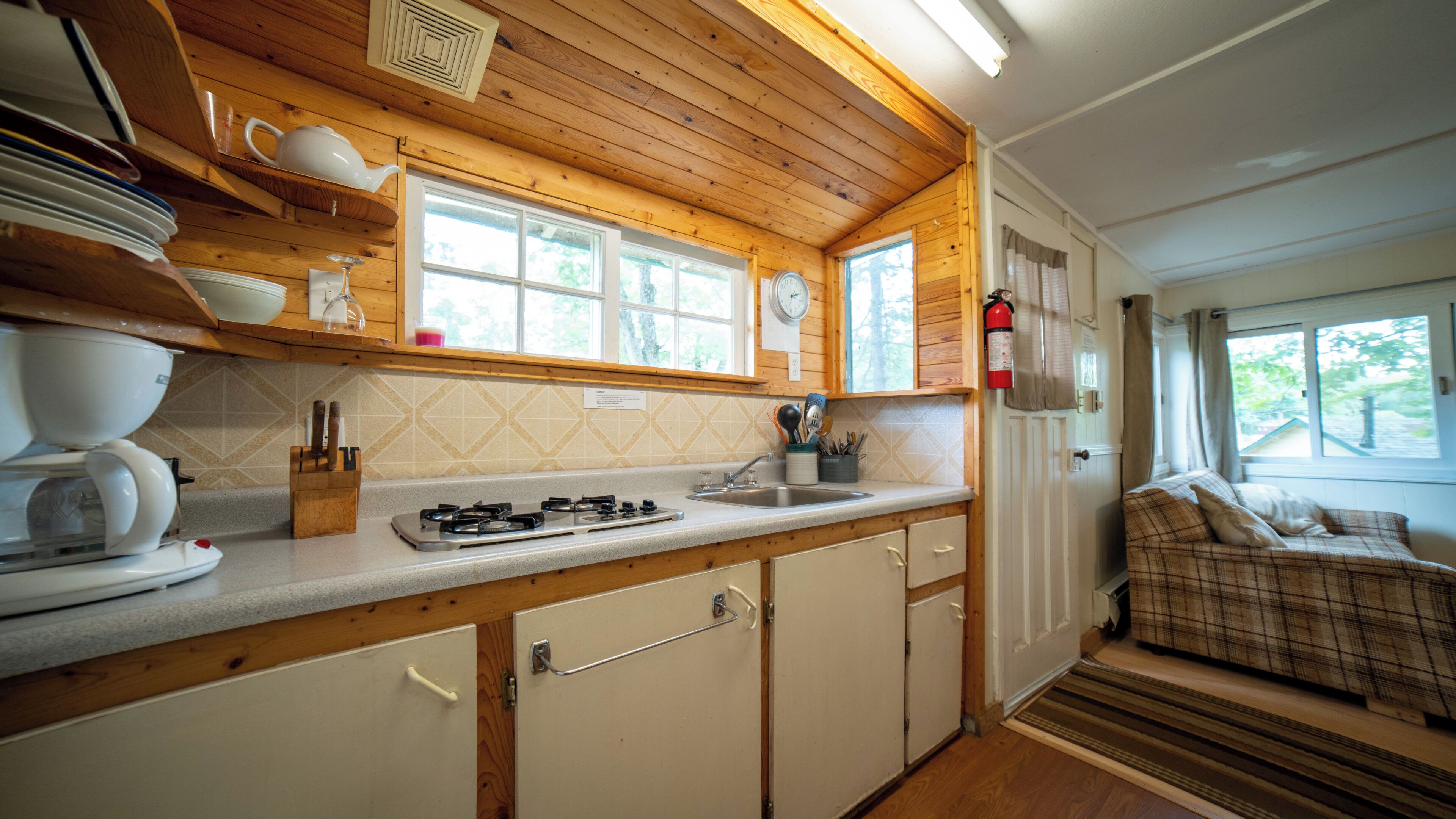 Eine kleine Küche mit Gasherd, Spüle und Holzschränken. Über der Spüle befindet sich ein Fenster und eine Tür führt in einen Wohnbereich mit einer Couch.