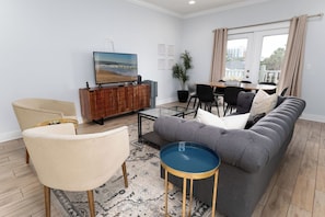 Ein Wohnzimmer mit Sofa, Sesseln, Couchtisch, Fernseher, Essbereich und einem großen Fenster.