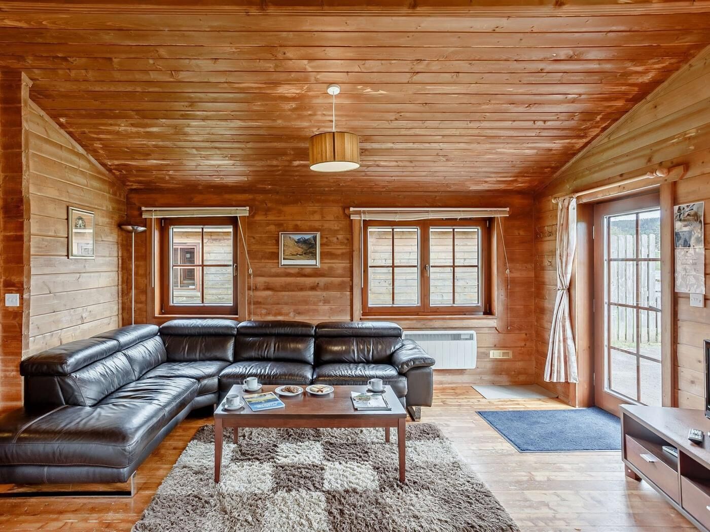 A log cabin interior with a leather sectional sofa, a wooden coffee table, and a hanging light fixture.