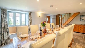 Un comedor con una mesa de madera dispuesta para seis personas, rodeada de sillas blancas, con una escalera y armarios de madera al fondo.