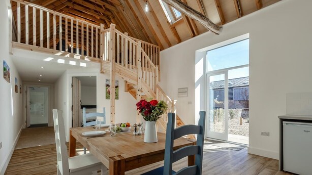 Un comedor amplio con una mesa de madera, sillas y un jarrón de flores. Una escalera conduce a un nivel superior con barandilla de madera.