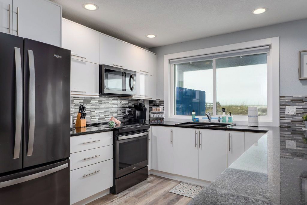 A modern kitchen with stainless steel appliances, white cabinetry, a large window, and a view of the outdoors.