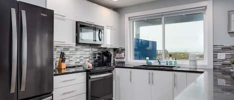 A modern kitchen with stainless steel appliances, white cabinetry, a large window, and a view of the outdoors.