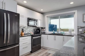A modern kitchen with stainless steel appliances, white cabinetry, a large window, and a view of the outdoors. - Front Bottom | Oceanfront steps from the sand! (Jacksonville Beach)