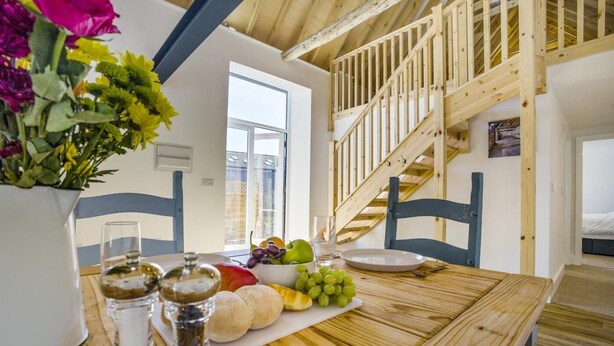 A wooden dining table with a fruit bowl, a vase with flowers, and salt and pepper shakers. A staircase with a wooden railing is visible in the background. - 1 Bed in Barrow (oc-b42641) (Barrow)