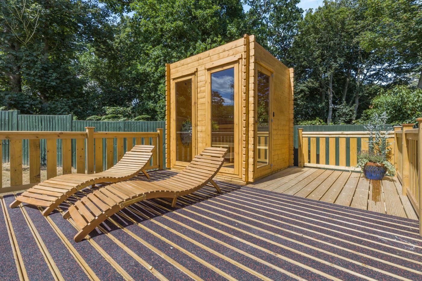A wooden sauna structure with two lounge chairs outside, surrounded by a wooden deck and a green fence.