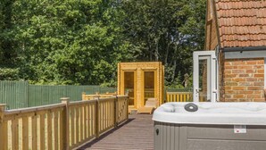 Une terrasse en bois mène à un espace de détente avec un jacuzzi et une porte en bois, le tout entouré d’une clôture verte et d’arbres.