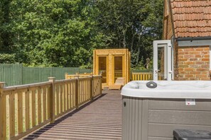 Eine Holzterrasse führt zu einem Wellnessbereich mit Whirlpool und Holztür, umgeben von einem grünen Zaun und Bäumen.