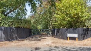 Une allée fermée par un portail mène à une propriété entourée d’une clôture en bois et agrémentée d’un banc.