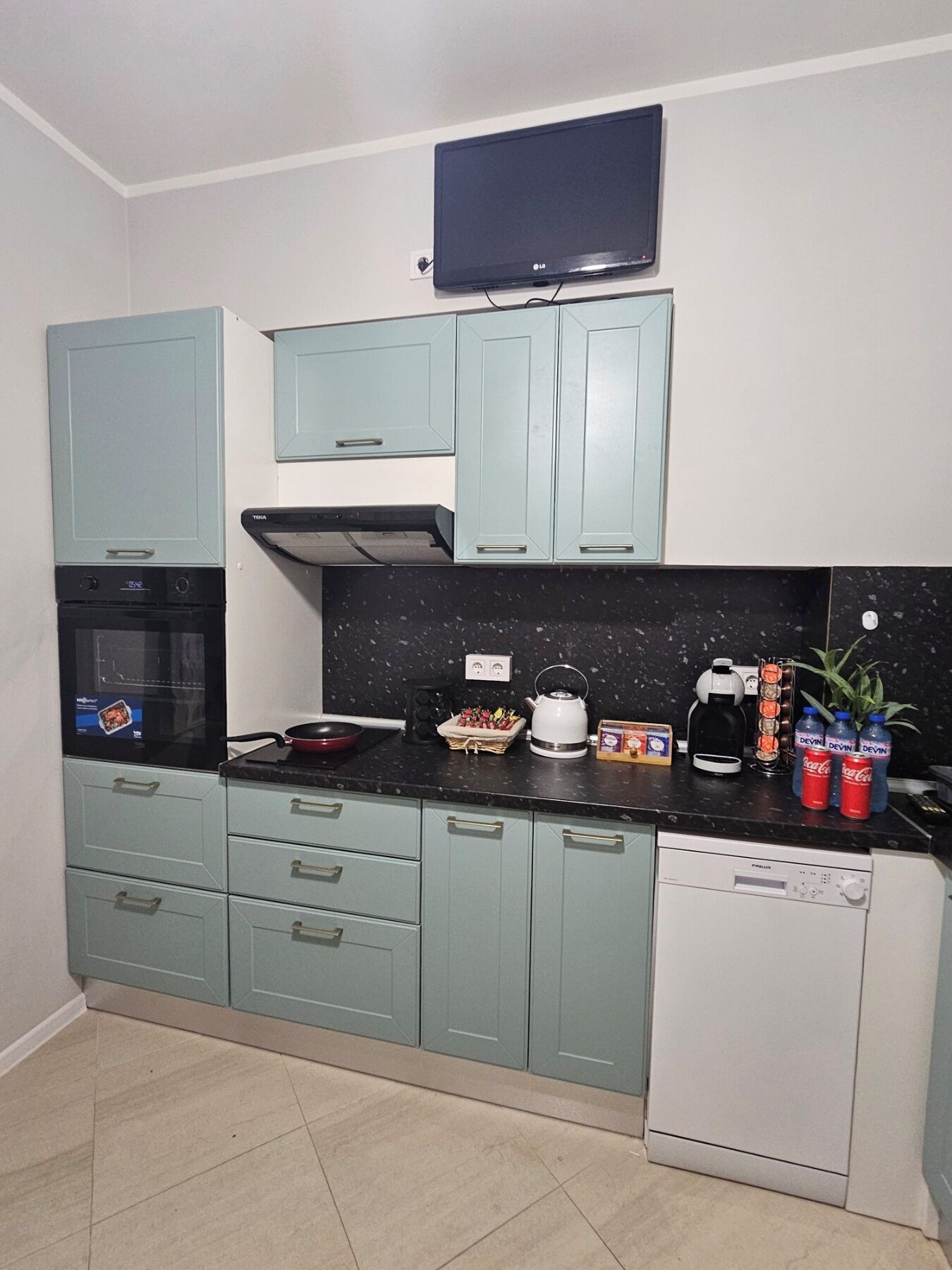 A compact kitchen with teal cabinets, a black countertop, a microwave, a kettle, and a dishwasher.