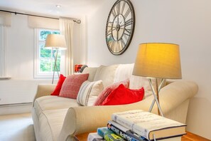 Ein Wohnzimmer mit einem beigen Sofa, roten und weißen Kissen mit Muster, einer gelben Lampe und einer Wanduhr.