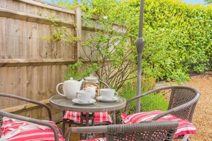A round table with a white teapot, cups, and saucers, surrounded by wicker chairs with red and white cushions.
