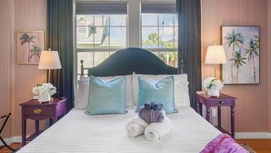 A hotel room with a bed, two nightstands, a window with curtains, and a view of buildings outside. - Fig Tree Inn | steps to the beach (Jacksonville Beach)
