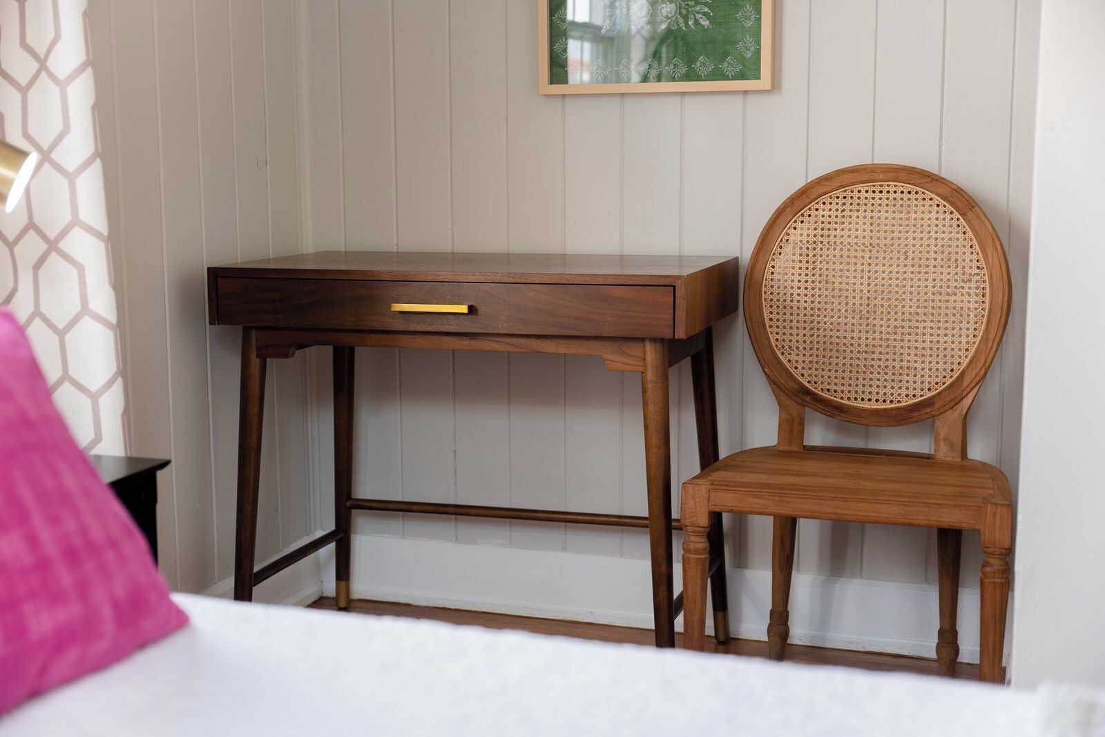 Una scrivania in legno con un cassetto e una sedia in rattan davanti a una parete bianca a pannelli.