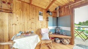 Interior acollidor d'una cabana de fusta, amb una taula preparada amb vasos i plats, una cadira, una estufa i vistes a l'exterior.
