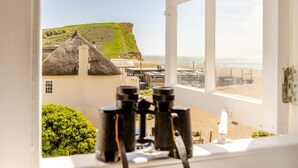 Ein Zimmer mit großem Fenster, das auf einen Strand Blick freigibt, ein Gebäude mit Reetdach und Fernglas auf dem Tisch.