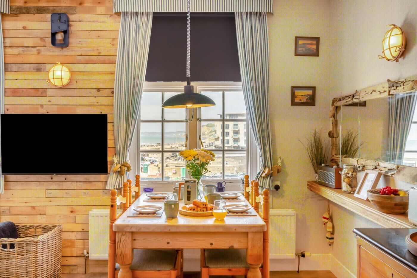 Une pièce chaleureuse avec un mur en bois, une table à manger dressée avec des assiettes et un vase de fleurs, une grande fenêtre avec des rideaux et un téléviseur à écran plat fixé au mur.