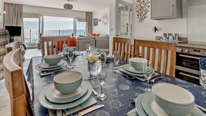 Zona pranzo con tavolo apparecchiato per quattro, cucina con elettrodomestici moderni e balcone con vista.