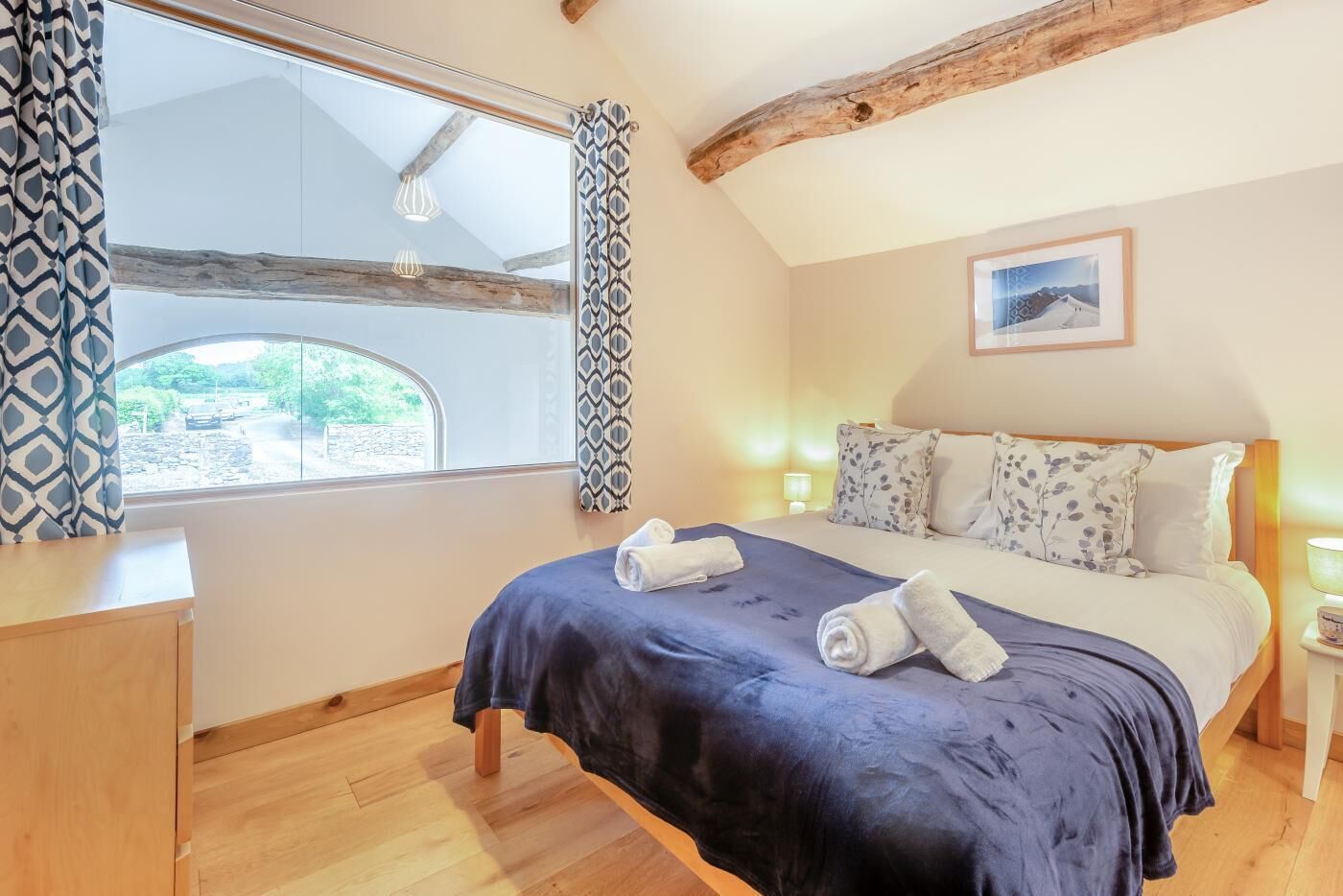 A bedroom with a bed, a window, a bedside table, and a wooden ceiling beam.